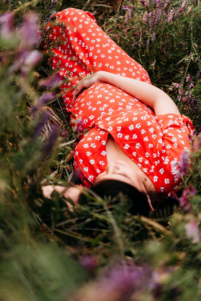 pregnant woman in a red dress with white flowers, cupping under her belly while laying in field of hairy purple vetch.  featured in post about the best time for maternity and newborn photos