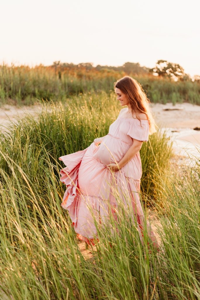 pregnant woman in a pink dress on a beach.  she is twirling the bottom of her dress with one hand, and cupping her baby bump with the other.  featured in post about the best time for maternity and newborn photos.